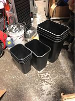 Nine black rectangular heavy duty garbage cans in three size groups, stacked and nested, visible in a workshop or storage area with other items.