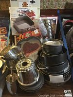 Photo depicting assorted baking pans including multiple springform pans, a metal bundt pan, a heart shaped glass baking pan, and metal flour sifters arranged on a table with boxed items in the background.