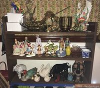 Wide view of shelving unit with assorted figurines and decorative items, including brass sculptures, porcelain human and bird figurines, and small dishes.