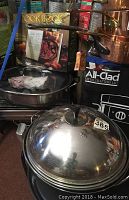 Photo shows stainless steel cookware stacked, copper-handled pot, and All-Clad branded electric grill and wok boxes in background with cookbooks.