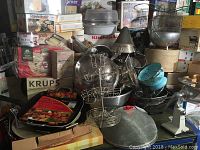 Kitchen utensils and small appliances stacked including Krups Sandwich Express box, strainers, and griddle.