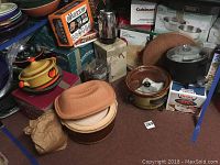 Photo shows assortment of kitchenware including crock pots in ceramic with lids, boxed cookware, coffee makers, and serving trays.