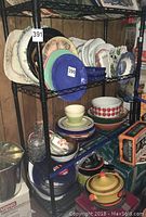 Entire black metal storage rack with four wire mesh shelves holding various plates and kitchen items.