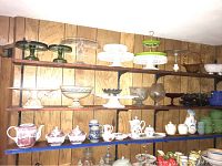 Wide view of wooden shelves filled with various glass cake stands, serving bowls, ceramic mugs and pitchers, and decorative glassware.