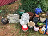 Wide view of assorted garden decor items including cement planters, ceramic pots, antique jug, arranged outdoors on ground.