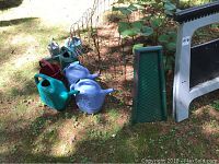 Items displayed outdoors including two folding saw horses, multiple watering cans in various colors, and a green garden kneeling pad on grass.
