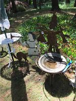 Outdoor image showing three metal garden decor sculptures: a white girl figure with an animal, a scarecrow figure, and a bird bath basin on stand.