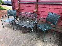 Two green metal outdoor chairs with woven straps and a green cast metal ornate bench placed against red siding outdoors.
