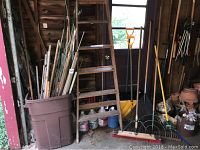 Overview of plant stakes in large brown container next to wooden ladder. Also visible are several yard tools including rake, snow shovel, garden fork, broom.