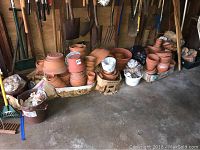 Multiple stacks and piles of terracotta garden pots, assorted sizes, with bags and containers of garden stones and beach shells, placed on concrete floor of garage area with garden tools visible in background.