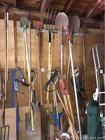 Photo of various yard and garden hand tools hung on a pegboard and wall rack inside a garage, including rakes, shovels, a hoe, and a post hole digger.