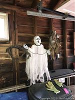 Photo showing hanging ghost figure with white tattered cloth and chain along with hanging scarecrow figure with ragged outfit in a garage setting.