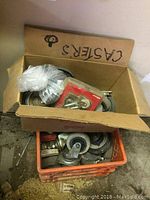 View inside open cardboard box and plastic crate packed with assorted casters showing various sizes and styles of caster wheels and mounting plates.