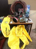 Photo shows wooden table with several items on top including spray guns, sandblasting kit hose and tools, buffer polisher, and yellow DuPont protective coveralls placed on and in front of the table.