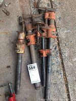 Four used pipe clamps arranged on concrete floor with MaxSold lot tag. Different pipe lengths visible; clamps painted orange with black or steel pipes.