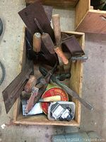 Top-down view of wooden box filled with assorted hand tools including trowels, hammer, clamps, and a tin of wire.