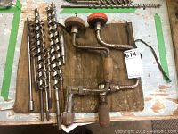 Photo shows two vintage hand drills and an assortment of drill bits placed on a worn wooden surface with green tape around the edges.
