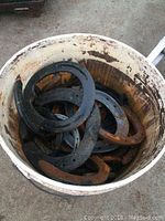 Bucket filled with various metal horse shoes showing rust and black coating in diverse sizes.