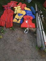 Overview of various life jackets in red, yellow, and blue alongside two sets of oars and a blue float, all placed on grass.