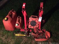 Overall view of red metal ladder jacks, two gas cans, ratchet strap and orange Noma cord caddy on grass.