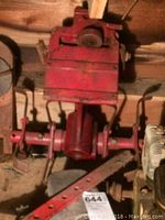 Red metal rotor tiller body with levers and gearbox, placed on the floor against a wall.