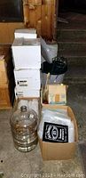 Cluster of wine making equipment boxes, large glass carboy, and accessories on floor in cellar or basement area.