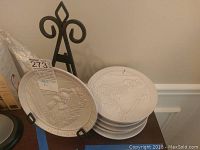 Stack of seven beige Frankoma collectible pottery plates with embossed designs and a decorative black metal display stand behind them.