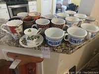 Eighteen assorted mugs displayed on kitchen countertop showing variety of designs and colors including holiday themes and blue and white scenic pattern.
