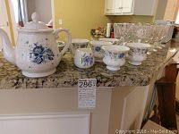 Full view of Royal Albert teapot, four cups (three Royal Albert, one Adderley), and crystal glassware on granite counter.