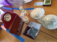 Photo showing all items in the lot on a table: two wrapped round Lenox holiday dishes, two ceramic snowmen candle holders, red wooden desk clock, wooden box holding pen, and two small ornaments in packaging.