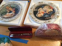 Photo showing two ceramic plates decorated with classical artwork and a red pen case with pen inside, with additional wrapped items.