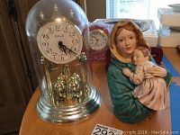 Anniversary clock with glass dome and gold-tone base alongside the Madonna statue on a wooden table under ambient daylight, showing clock face and pendulum details.