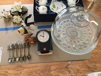 Overview of cake stand with dome, travel clock, silver spoons, and ceramic rooster figurine