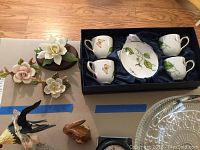 Overview of lot showing boxed set of 4 cups and saucers with butterflies and leaves design, and 3 porcelain hand-painted flower figurines on table.