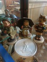 Four collectible Hummel figurines displayed together including a boy with a green hat, a girl with umbrella, and others, featuring detailed hand-painted porcelain finish.