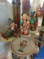 Three Hummel figurines displayed on glass shelf showing a reclining violin player, standing cello player, and double bass player.