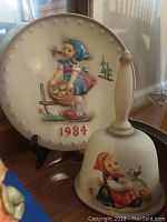 Plate with raised illustration of a girl and 1984 year marking, accompanied by a decorative bell featuring a girl and bird.