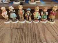 Front view of 7 Hummel figurines in a row on wood floor, showing detailed colorful clothing and poses.
