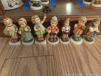 Front view of all 7 Hummel figurines standing on wooden floor with wood furniture behind