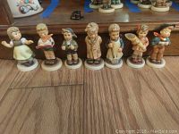 Front view of seven Hummel figurines in a row on a wooden floor, showcasing their detailed designs and colors.