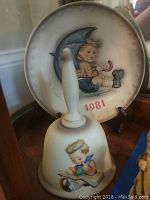 Porcelain 1981 Hummel plate with illustration of a child under a moon and the year 1981 printed on it, accompanied by a 1980 Hummel bell with a child reading a book illustration.