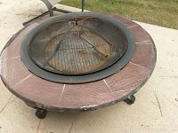 Top and angle view showing round fire pit with reddish stone-like border, mesh cover with handle, and curled metal legs on patio surface.