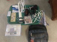 Top view of all items on a green tray including two compact hair dryers, handheld massager, Omron blood pressure monitor in box, power strip, black carrying bag, and other small personal care items.