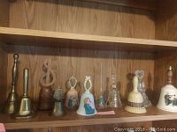 Shelf display with eleven assorted bells made from brass, ceramic, and crystal in varying sizes and styles.