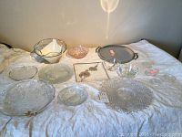 Displayed on white cloth, assortment of glass platters, bowls, and Danbury Mint paperweights showing varying etched and pressed glass designs and shapes