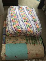 Photo of folded handmade quilt at bottom with multicolor crochet afghan folded on top, showing patchwork squares of different colors and patterns.