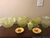 Photo showing four yellow hobnail glass pieces and two yellow ceramic flower-shaped tea bag holders arranged on a wooden surface.