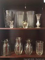 Two shelves of clear glass vases, containers, and paperweight, showing variety in design including ribbed and cut patterns and one metal stemmed apple paperweight.
