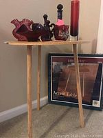 Full view of a wood accent table with round top holding several red glass collectible items including a bell and vases, the original box visible behind.