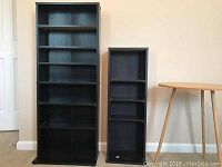 Two black CD racks standing side by side against wall with a round wooden three-leg table on the right.